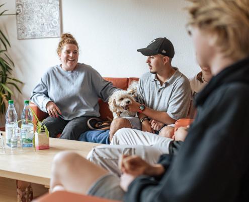 Pädagogische Fachkraft sitzt gemeinsam mit Jugendlichen und einem Hund auf Sofas einer Internatsgruppe